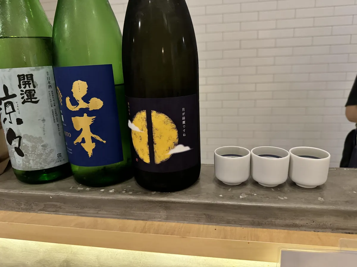 カウンターに並ぶ日本酒のボトルと、三つのお猪口。右端に「つきが綺麗ですね」のボトルが見える
